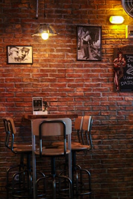 A cozy bar interior with a brick wall adorned with vintage black-and-white photographs and a retro-style chalkboard sign. A small wooden table is surrounded by four high metal chairs. Soft, warm lighting from decorative lamps enhances the intimate atmosphere.