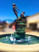 A statue depicts a girl and a boy on a stack of books. The girl is joyfully leaning backward with her arms spread wide, while the boy is seated, focusing on a book. The sculpture sits in the center of a circular fountain with water jets.