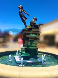A statue depicts a girl and a boy on a stack of books. The girl is joyfully leaning backward with her arms spread wide, while the boy is seated, focusing on a book. The sculpture sits in the center of a circular fountain with water jets.