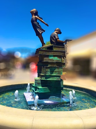 A statue depicts a girl and a boy on a stack of books. The girl is joyfully leaning backward with her arms spread wide, while the boy is seated, focusing on a book. The sculpture sits in the center of a circular fountain with water jets.