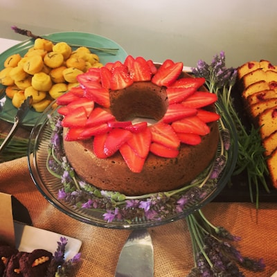 A beautifully arranged table features a large chocolate cake topped with sliced strawberries, surrounded by sprigs of lavender. To the left, a plate of small yellow pastry puffs is visible. On the right, slices of a loaf cake are presented, with sprigs of greenery and a rustic cloth enhancing the presentation.