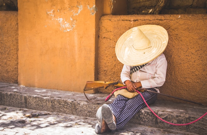 A person wearing a large straw hat and traditional clothing is sitting against an adobe wall on a stone step, holding a wooden musical instrument or a tool with a pink cord.