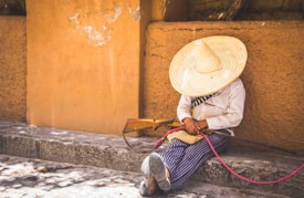 A person wearing a large straw hat and traditional clothing is sitting against an adobe wall on a stone step, holding a wooden musical instrument or a tool with a pink cord.
