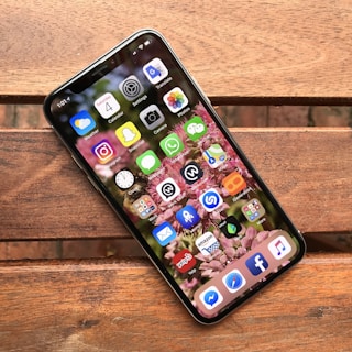 A smartphone is placed on a wooden surface, displaying its home screen filled with various app icons such as Instagram, Snapchat, and Facebook, with a floral wallpaper background.