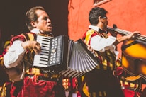 Two musicians wearing traditional, colorful costumes are playing instruments. One musician is playing an accordion, while the other is playing a double bass. They are performing energetically in front of a vivid red wall.