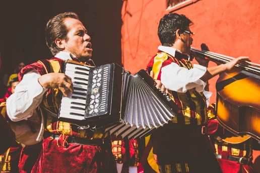 Two musicians wearing traditional, colorful costumes are playing instruments. One musician is playing an accordion, while the other is playing a double bass. They are performing energetically in front of a vivid red wall.
