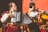 Two musicians wearing traditional, colorful costumes are playing instruments. One musician is playing an accordion, while the other is playing a double bass. They are performing energetically in front of a vivid red wall.
