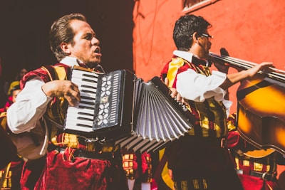 Two musicians wearing traditional, colorful costumes are playing instruments. One musician is playing an accordion, while the other is playing a double bass. They are performing energetically in front of a vivid red wall.