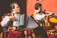 Two musicians wearing traditional, colorful costumes are playing instruments. One musician is playing an accordion, while the other is playing a double bass. They are performing energetically in front of a vivid red wall.