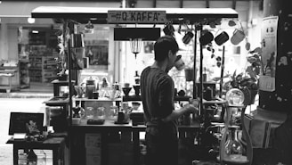 A person is standing at a small, street-side coffee stall, engaged in making a beverage. The stall is equipped with various coffee-making tools and decorations, including hanging mugs, pots, and small plants. The atmosphere suggests a quaint and cozy environment.