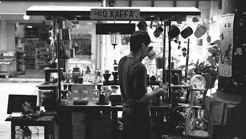 A person is standing at a small, street-side coffee stall, engaged in making a beverage. The stall is equipped with various coffee-making tools and decorations, including hanging mugs, pots, and small plants. The atmosphere suggests a quaint and cozy environment.
