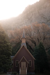 A quaint brown wooden chapel with a steeply pitched roof and a small belfry stands surrounded by dense trees. The backdrop features towering rocky cliffs, and bright sunlight diffuses over the scene, creating a serene and tranquil atmosphere.