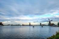 A serene landscape featuring traditional Dutch windmills beside a calm body of water. The sky is filled with scattered clouds, creating a peaceful and picturesque scene. Several buildings with red roofs are visible near the windmills, adding a charming touch to the rural setting.