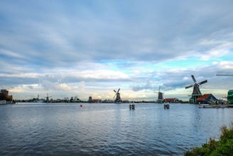 A serene landscape featuring traditional Dutch windmills beside a calm body of water. The sky is filled with scattered clouds, creating a peaceful and picturesque scene. Several buildings with red roofs are visible near the windmills, adding a charming touch to the rural setting.