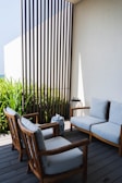 A cozy outdoor seating area features wooden chairs and a couch with light gray cushions, arranged on a wooden deck. Tall, green plants grow behind a decorative vertical slatted screen. A small blue and white decorative jar and a metal lantern are placed next to the seating.