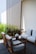 A cozy outdoor seating area features wooden chairs and a couch with light gray cushions, arranged on a wooden deck. Tall, green plants grow behind a decorative vertical slatted screen. A small blue and white decorative jar and a metal lantern are placed next to the seating.