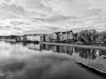 A serene riverside scene with a row of traditional European buildings lining the waterfront. The calm water reflects the buildings and sky, creating a mirror effect. Leafy trees add a natural element, while the sky is partly cloudy with hints of sunlight peeking through.
