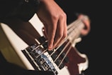 A close-up view of a hand playing a bass guitar, focusing on the fingers plucking the strings. The background is dark, highlighting the instrument and the player's hand.