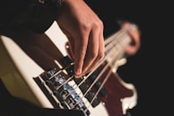 A close-up view of a hand playing a bass guitar, focusing on the fingers plucking the strings. The background is dark, highlighting the instrument and the player's hand.