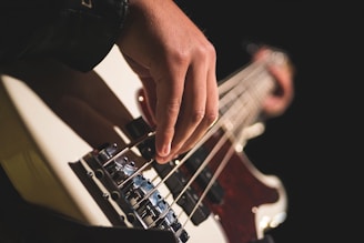 A close-up view of a hand playing a bass guitar, focusing on the fingers plucking the strings. The background is dark, highlighting the instrument and the player's hand.