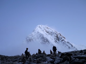 A majestic, snow-covered mountain peak rises against a clear blue sky. In the foreground, several hikers are walking on rocky terrain, silhouetted by the light. Mist and clouds partially obscure the lower part of the mountain, creating a mysterious and captivating atmosphere.