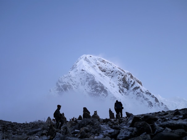 A majestic, snow-covered mountain peak rises against a clear blue sky. In the foreground, several hikers are walking on rocky terrain, silhouetted by the light. Mist and clouds partially obscure the lower part of the mountain, creating a mysterious and captivating atmosphere.