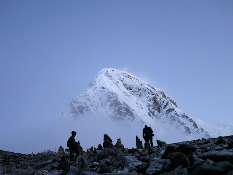 A majestic, snow-covered mountain peak rises against a clear blue sky. In the foreground, several hikers are walking on rocky terrain, silhouetted by the light. Mist and clouds partially obscure the lower part of the mountain, creating a mysterious and captivating atmosphere.