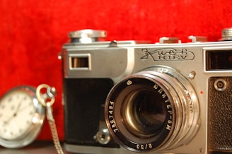 A vintage camera is prominently displayed against a vibrant red background. The camera, with a metallic body and detailed engravings, is styled in a classic design. To the side, there is a pocket watch with a silver chain, slightly blurred, adding a nostalgic feel.