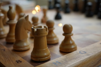 Several light brown wooden chess pieces are positioned on a chessboard, with a rook in the foreground and other pieces like a knight and pawns nearby. The surface of the board is marked with alphanumeric coordinates. Soft lighting creates a warm atmosphere, with fairy lights shimmering in the background.