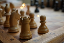 Several light brown wooden chess pieces are positioned on a chessboard, with a rook in the foreground and other pieces like a knight and pawns nearby. The surface of the board is marked with alphanumeric coordinates. Soft lighting creates a warm atmosphere, with fairy lights shimmering in the background.