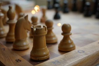 Several light brown wooden chess pieces are positioned on a chessboard, with a rook in the foreground and other pieces like a knight and pawns nearby. The surface of the board is marked with alphanumeric coordinates. Soft lighting creates a warm atmosphere, with fairy lights shimmering in the background.