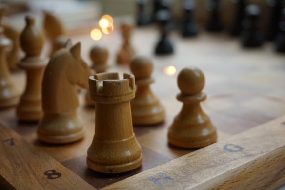 Several light brown wooden chess pieces are positioned on a chessboard, with a rook in the foreground and other pieces like a knight and pawns nearby. The surface of the board is marked with alphanumeric coordinates. Soft lighting creates a warm atmosphere, with fairy lights shimmering in the background.