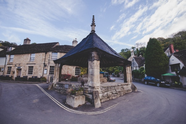 A picturesque village scene with historic stone buildings lining a quiet street. The focal point is a roofed stone structure with a quaint, rustic appearance. Surrounding the buildings are lush trees and greenery, indicating a rural setting. Cars are parked along the street, suggesting a peaceful and charming countryside atmosphere.