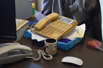 A cluttered desk with an old-fashioned beige telephone featuring a keypad and display screen. Next to it is another phone handset, a mug with a brown design, a stack of paper, a screwdriver with a red handle, and a boxed item partially visible. The background includes a computer monitor, glue stick, and various office supplies.