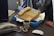 A cluttered desk with an old-fashioned beige telephone featuring a keypad and display screen. Next to it is another phone handset, a mug with a brown design, a stack of paper, a screwdriver with a red handle, and a boxed item partially visible. The background includes a computer monitor, glue stick, and various office supplies.