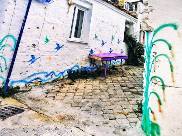A quaint, cobblestone alley is adorned with vibrant street art, featuring colorful birds in flight and playful waves painted on whitewashed walls. A bright purple table adds a bold pop of color to the charming, rustic setting.