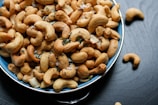 A blue bowl filled with roasted cashew nuts, garnished with coarse salt and small green herbs, placed on a dark surface with a few cashews scattered around the bowl.