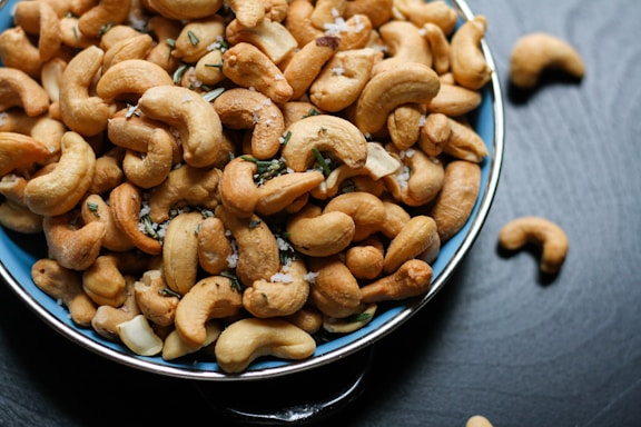 A blue bowl filled with roasted cashew nuts, garnished with coarse salt and small green herbs, placed on a dark surface with a few cashews scattered around the bowl.