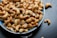 A blue bowl filled with roasted cashew nuts, garnished with coarse salt and small green herbs, placed on a dark surface with a few cashews scattered around the bowl.