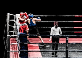 A boxing match in progress features two fighters inside a ring, one wearing red gear and the other in blue. The referee stands close by, ensuring the match proceeds fairly. The ring is equipped with ropes and padded corners, set against a black background, highlighting the intense action taking place.