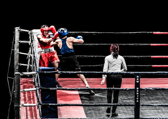 A boxing match in progress features two fighters inside a ring, one wearing red gear and the other in blue. The referee stands close by, ensuring the match proceeds fairly. The ring is equipped with ropes and padded corners, set against a black background, highlighting the intense action taking place.