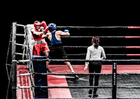 A boxing match in progress features two fighters inside a ring, one wearing red gear and the other in blue. The referee stands close by, ensuring the match proceeds fairly. The ring is equipped with ropes and padded corners, set against a black background, highlighting the intense action taking place.