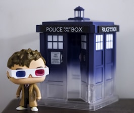 A figurine of a person wearing 3D glasses and a brown coat stands next to a small model of a blue police box, featuring the words 'POLICE PUBLIC CALL BOX' at the top.