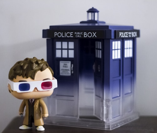 A figurine of a person wearing 3D glasses and a brown coat stands next to a small model of a blue police box, featuring the words 'POLICE PUBLIC CALL BOX' at the top.