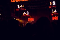 The image captures a dark concert or festival setting featuring illuminated screens displaying the text 'Alta Voz 2017'. The scene includes stage equipment and what appears to be a large outdoor event with colorful lighting effects.