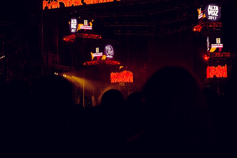 The image captures a dark concert or festival setting featuring illuminated screens displaying the text 'Alta Voz 2017'. The scene includes stage equipment and what appears to be a large outdoor event with colorful lighting effects.