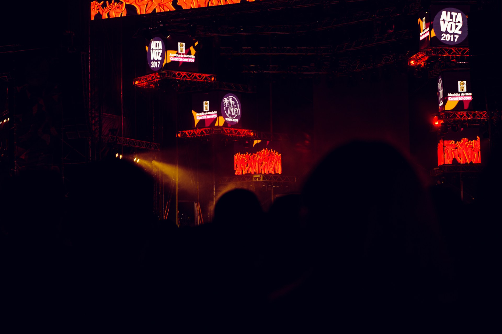 The image captures a dark concert or festival setting featuring illuminated screens displaying the text 'Alta Voz 2017'. The scene includes stage equipment and what appears to be a large outdoor event with colorful lighting effects.