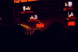 The image captures a dark concert or festival setting featuring illuminated screens displaying the text 'Alta Voz 2017'. The scene includes stage equipment and what appears to be a large outdoor event with colorful lighting effects.