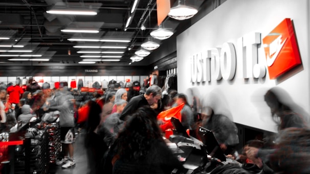 A busy retail store with numerous people examining products and clothing. The store appears to be selling athletic wear, with the phrase 'Just Do It.' prominently displayed on a wall alongside a recognizable orange logo. The interior is well-lit with modern lighting, shelves filled with merchandise, and a counter where people are gathered, possibly checking out or asking questions.