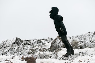 A person dressed in heavy winter clothing stands on a rocky, snow-covered terrain. They are wearing a hooded jacket and boots, appearing to brace against the cold, with a small camera hanging by their side. The landscape is dominated by rugged rocks and a light dusting of snow, under a gray sky.